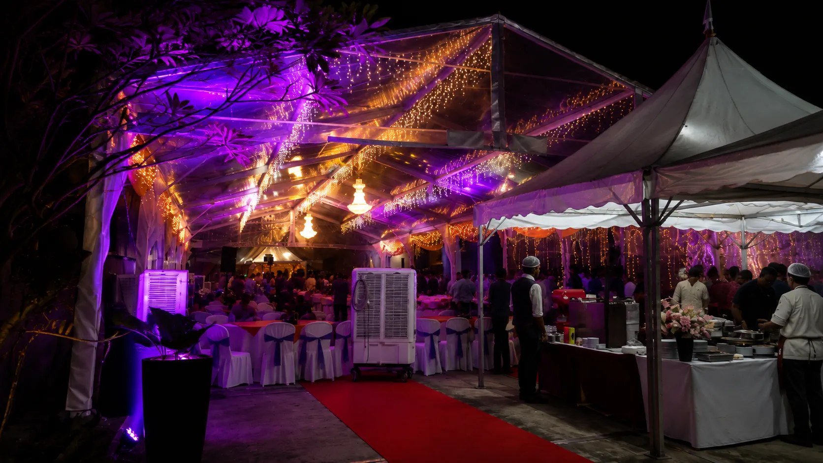 Clear roof wedding banquet marquee at night with red carpet walkway, chandelier lighting, purple uplighting, dining tables and buffet service tent